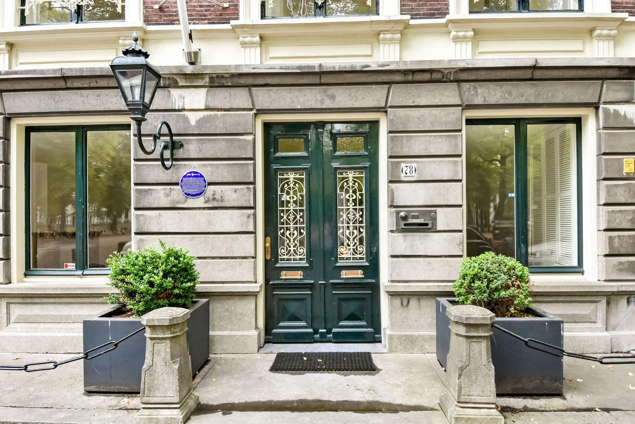 Ingang van een historisch pand aan het Lange Voorhout 28 in Den Haag, met twee sierlijke groene deuren, een blauwe herdenkingsplaquette en symmetrische plantenbakken.